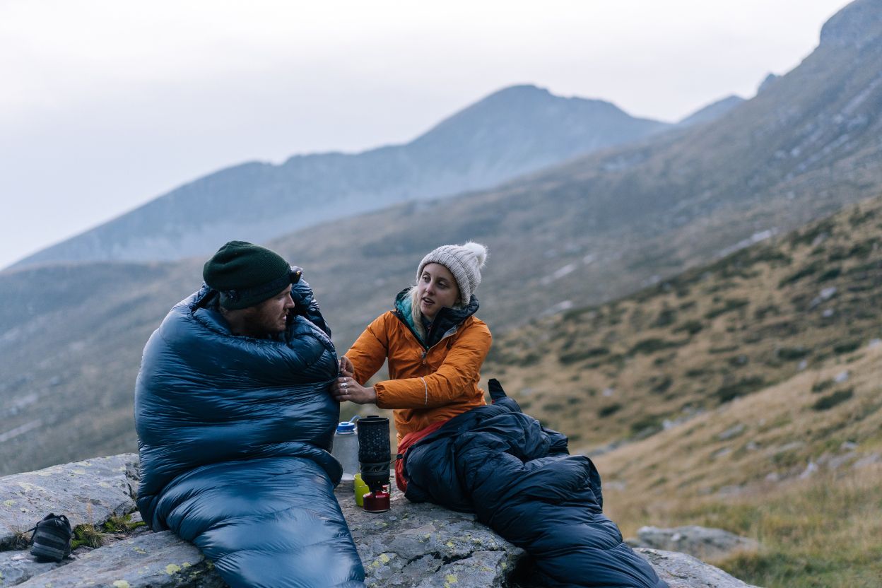 A man and woman wrapped up in their sleeping bags, while they wait on the rest of their group.