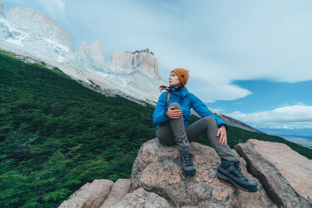 A woman hiker sitting on a rock, mountain massif and forest behind.