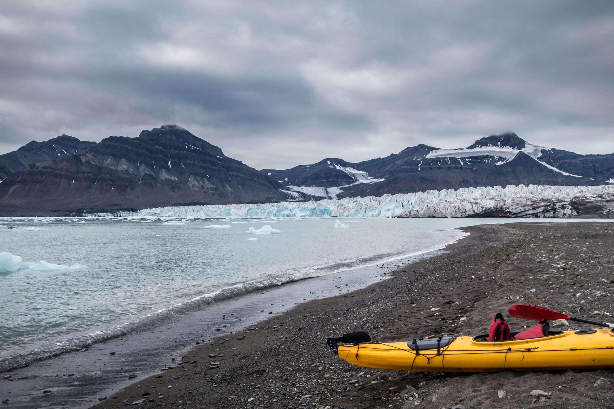 A kayak on a dark beach in Svalbard, with glaciers and mountains in the background.