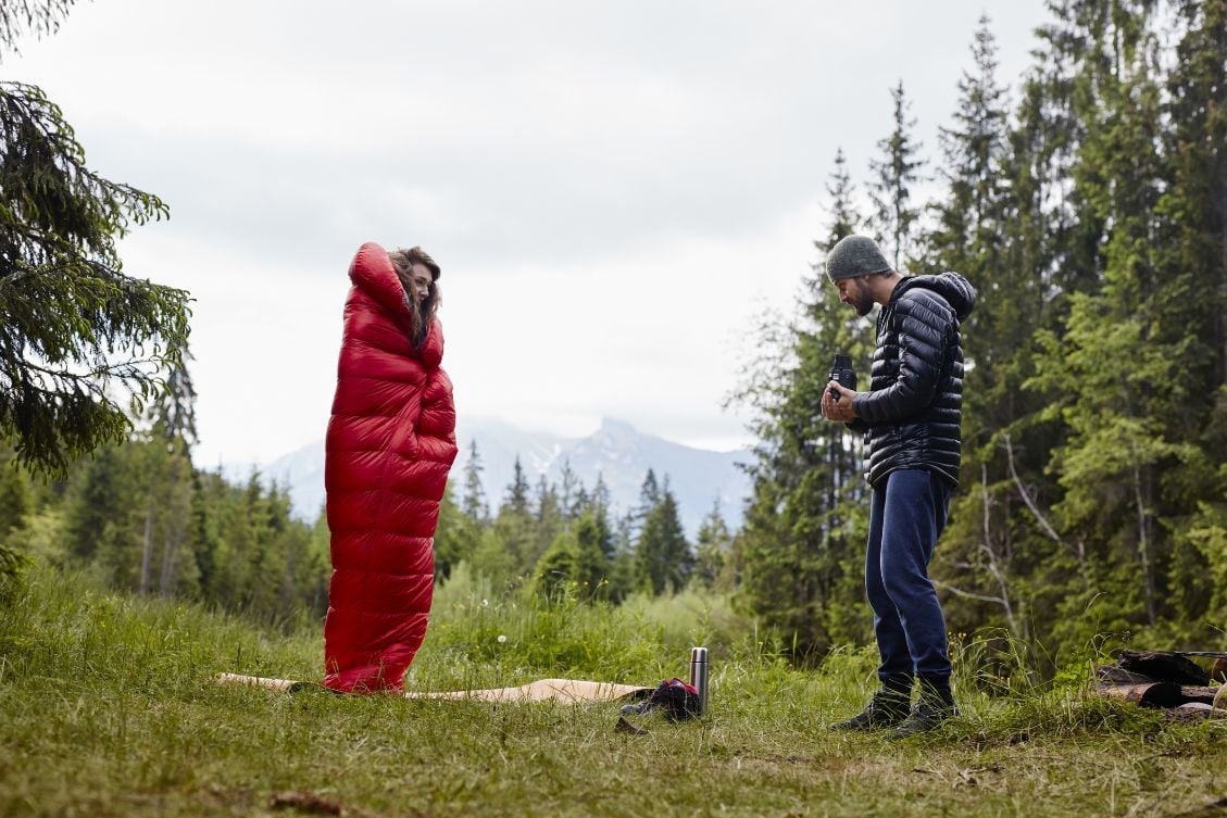 A woman wrapped up in a sleeping bag, while a man takes a photo in front of a beautiful view.