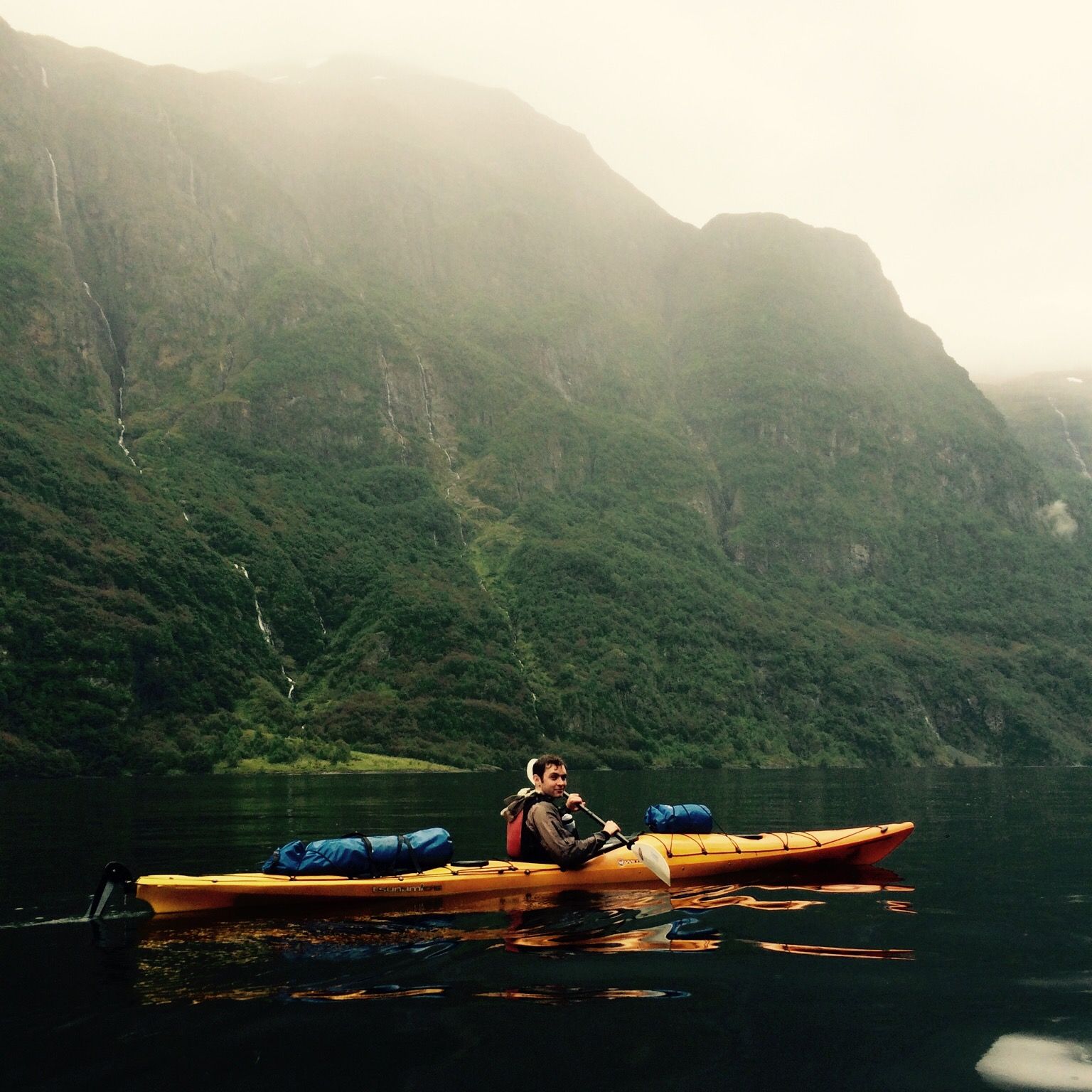 A man kayaks, with a backdrop of the dark green and wet landscape of the Norwegian fjords