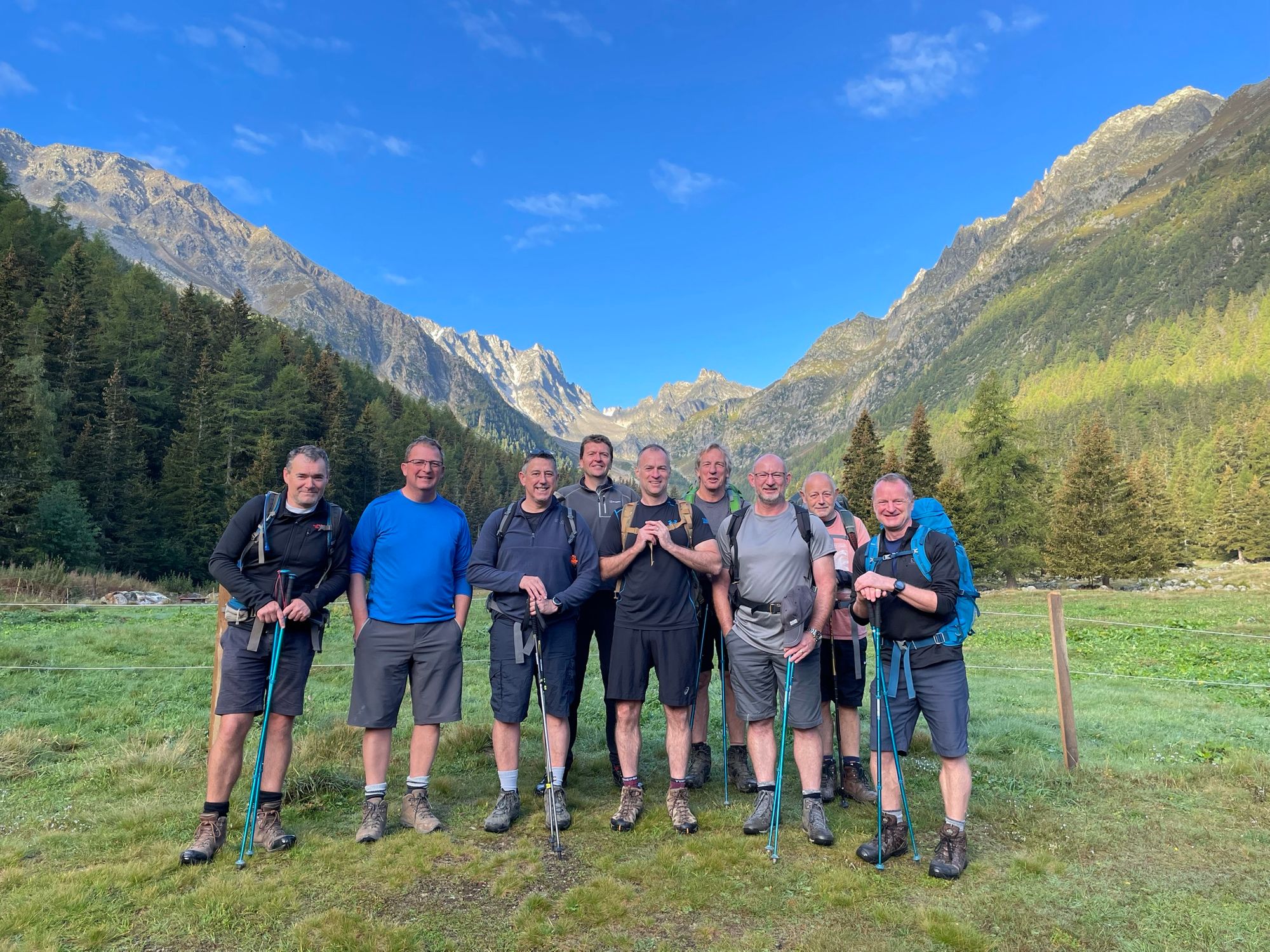 A group of hikers poses for a photo in an Alpine meadow, with mountains behind.