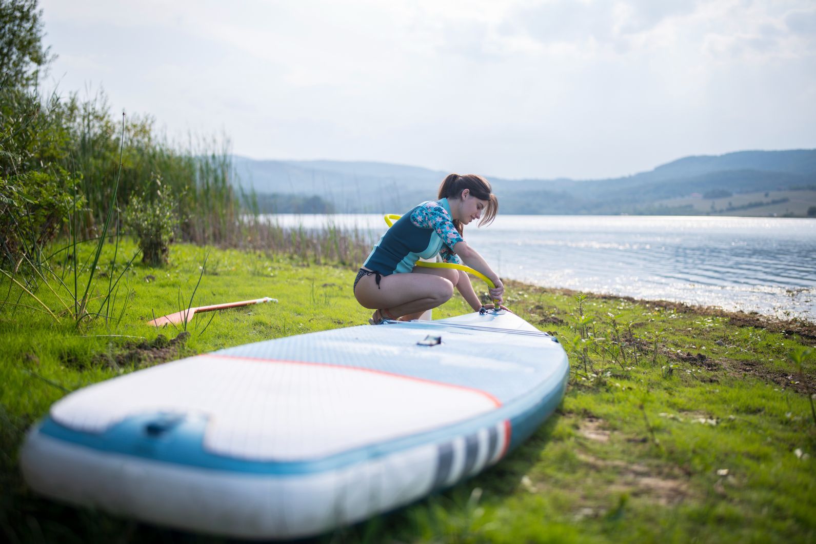 A woman inflates her stand-up paddleboard on the edge of a lake.