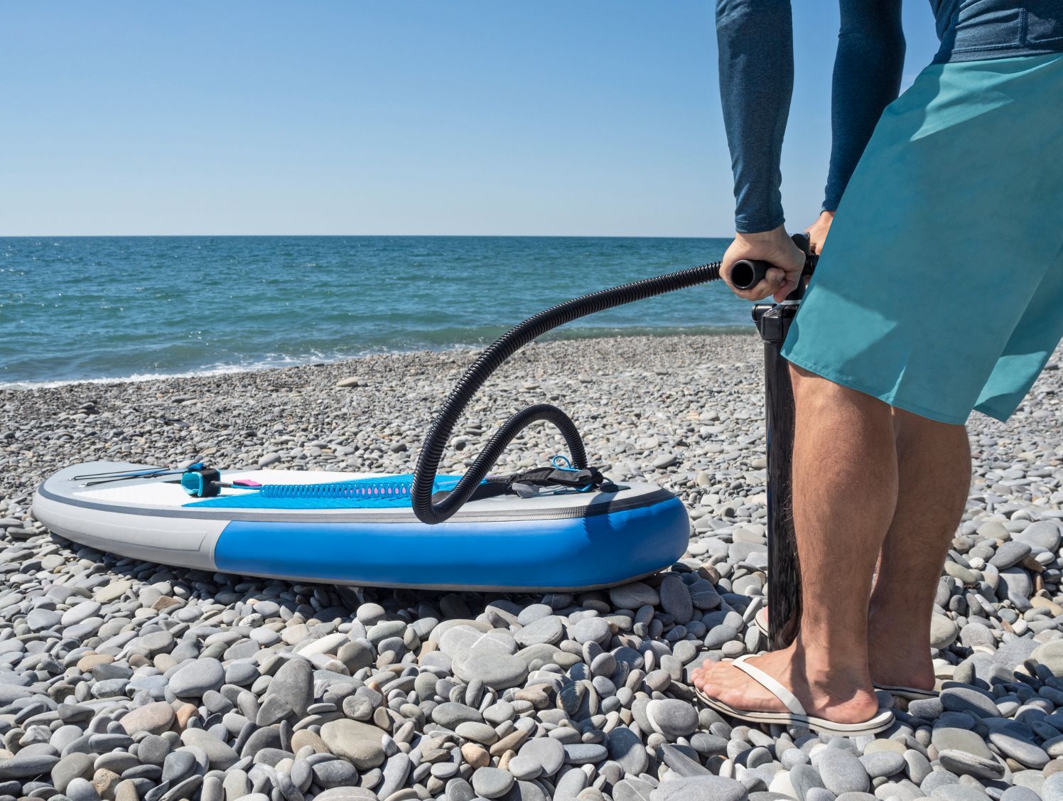 A person in flip flops pumps up a stand-up paddleboard on a rocky beach, next to the water.