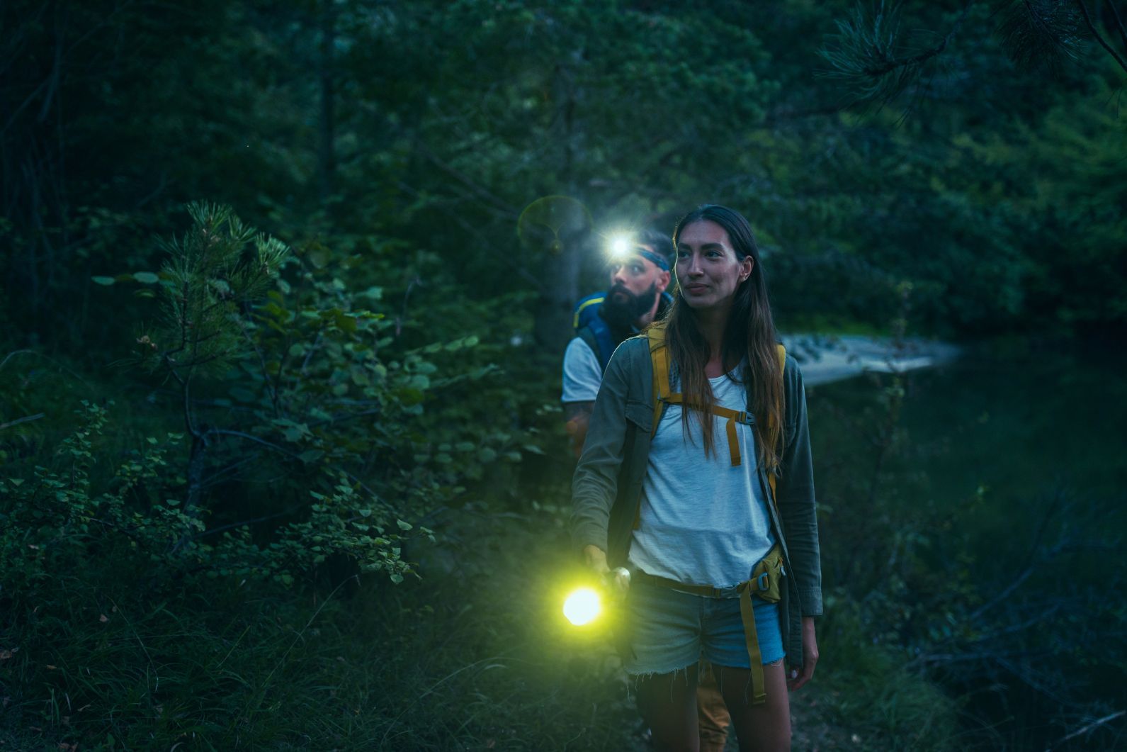 Two hikers at night in the forest, one with a torch and another with a headlamp, looking scared.