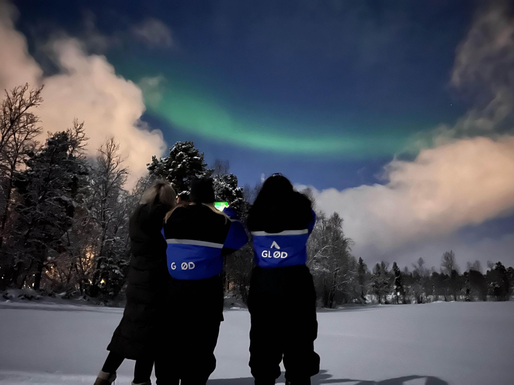 People photograph the northern lights from the shores of a frozen lake.