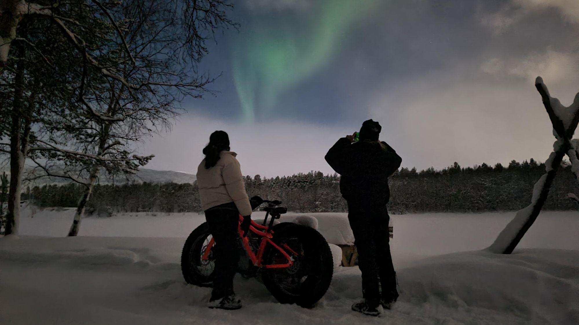 The northern lights blaze in the sky as two people watch, fat bikes by their sides.