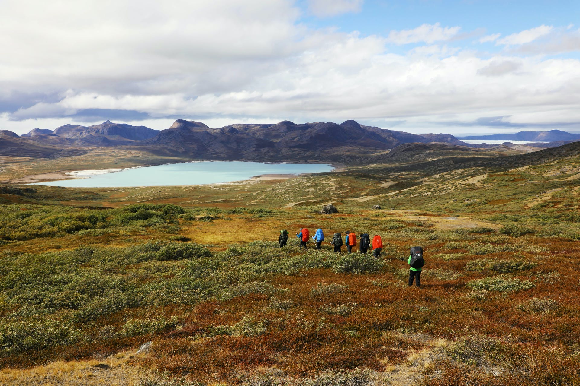 A group of hikers treks across the Greenland wilderness - a lake and mountains in the background.