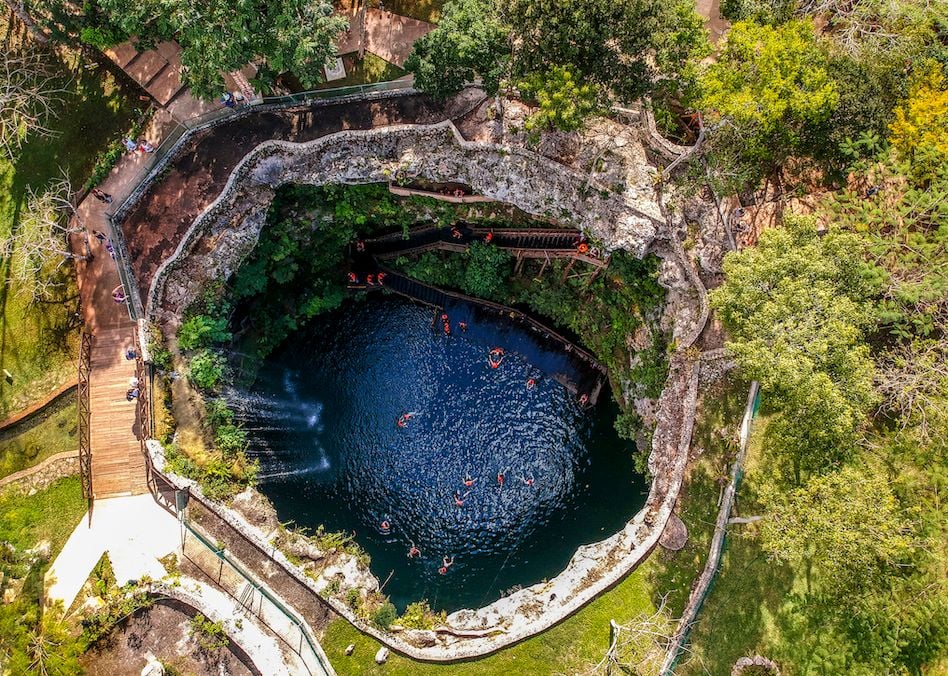 A view of a cenote seen from above, with tourists swimming in its blue waters