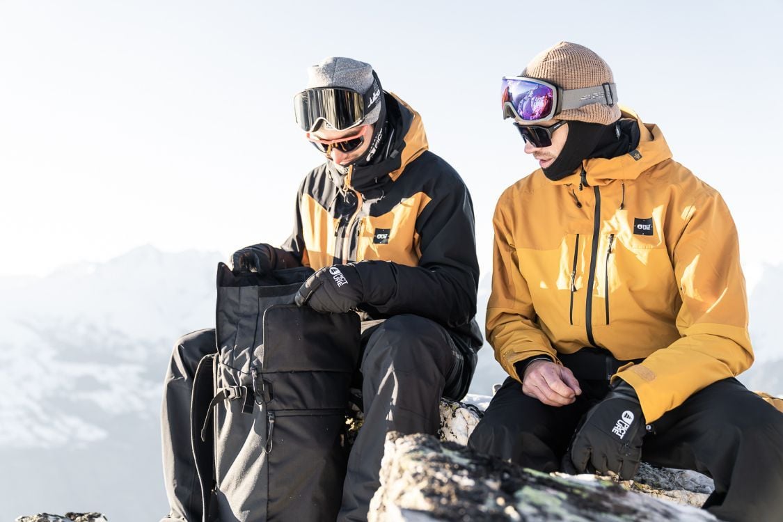 Two skiers wearing Picture Organic rental clothing dig into a rucksack on a mountain top.