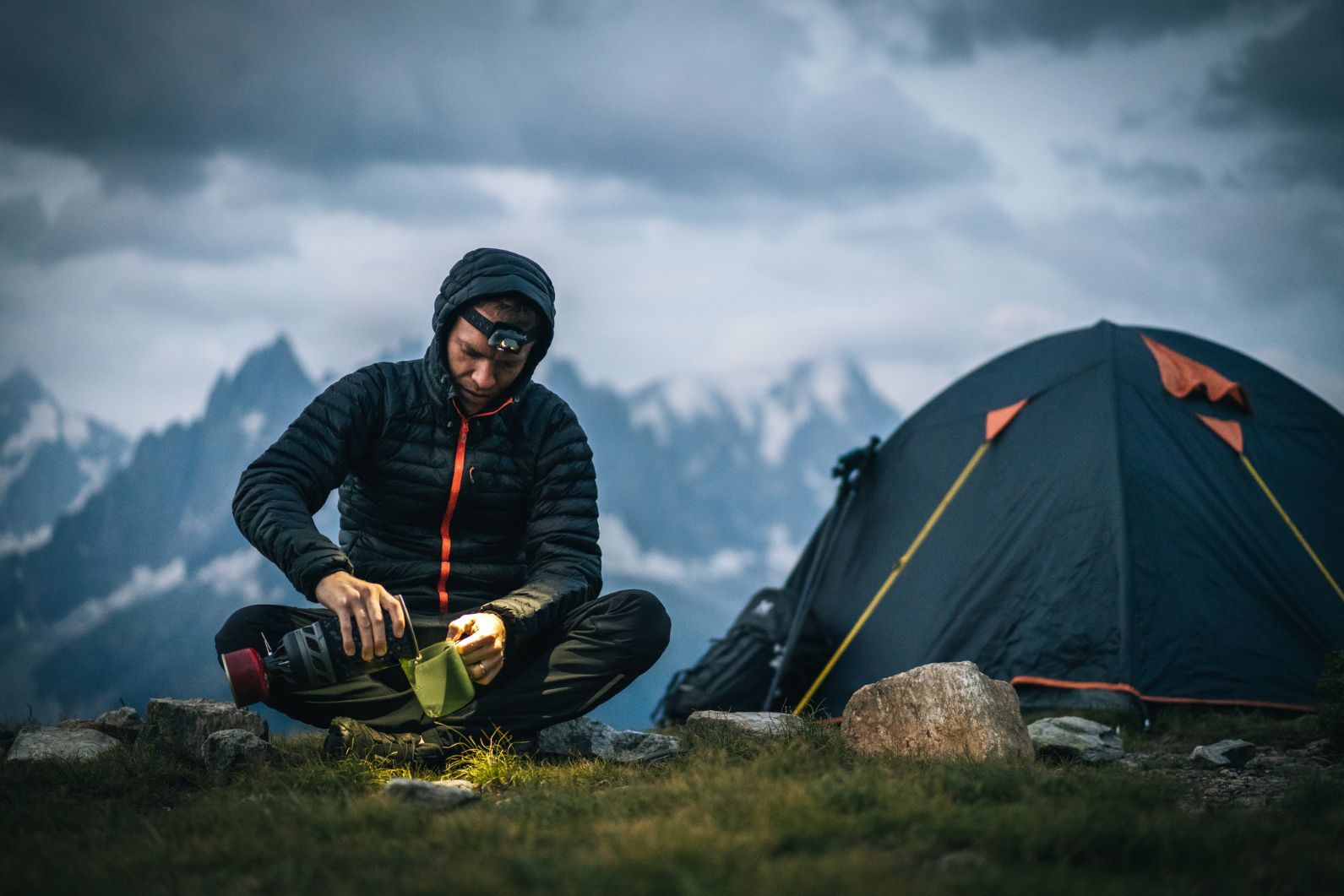 A camper fills up a mug with boiling water made over a camping stove, next to his tent, with a backdrop of mountains.