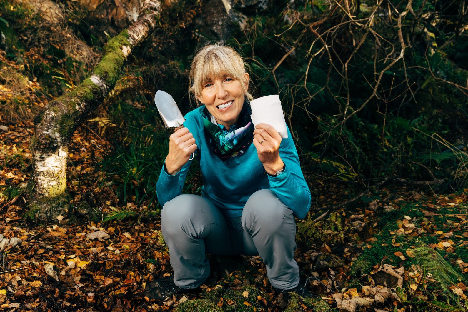 A woman squats down to go the toilet, with a trowel and toilet roll.