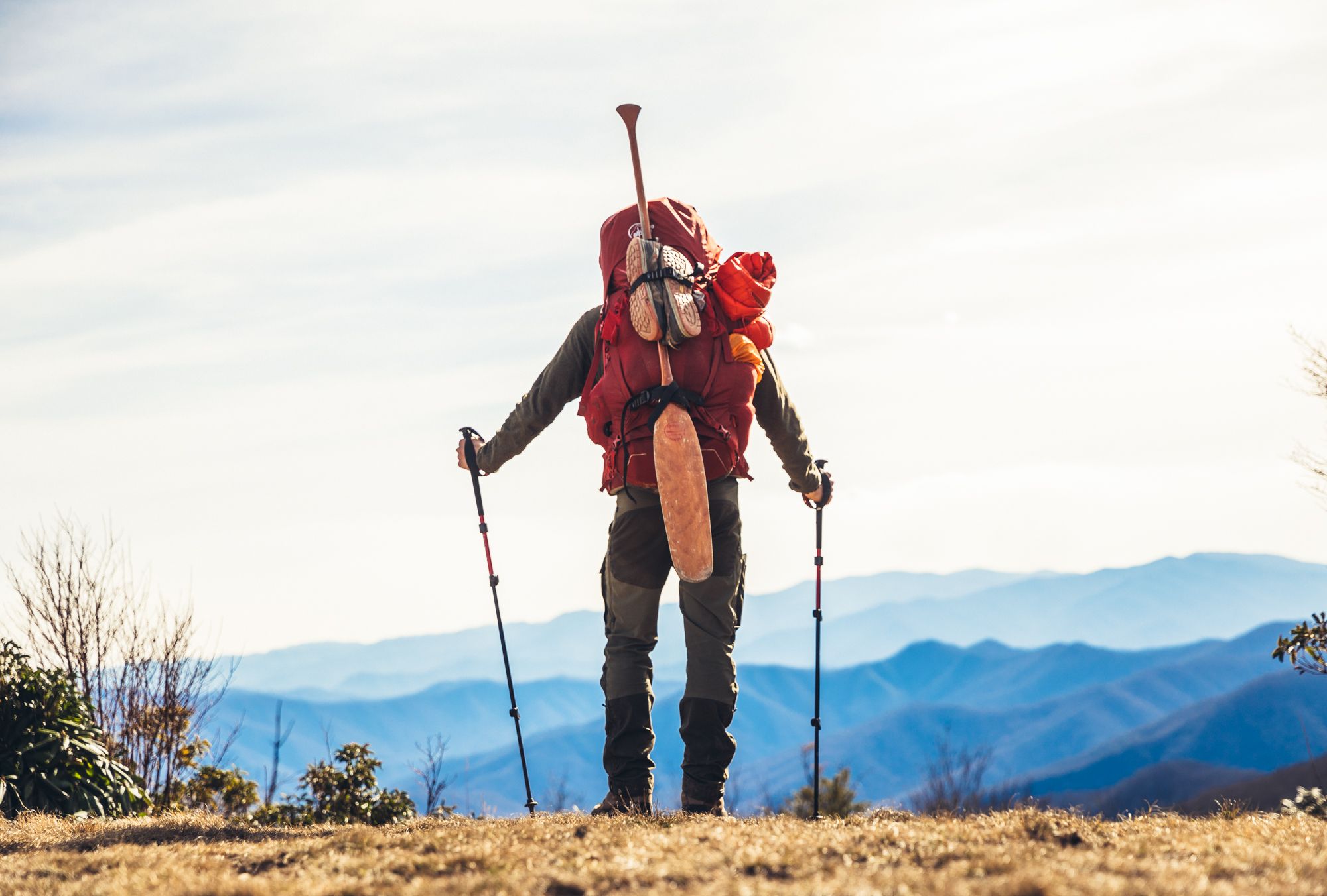 Hiker facing towards the mountains. A full backpack has an oar and shoes strapped to the back.