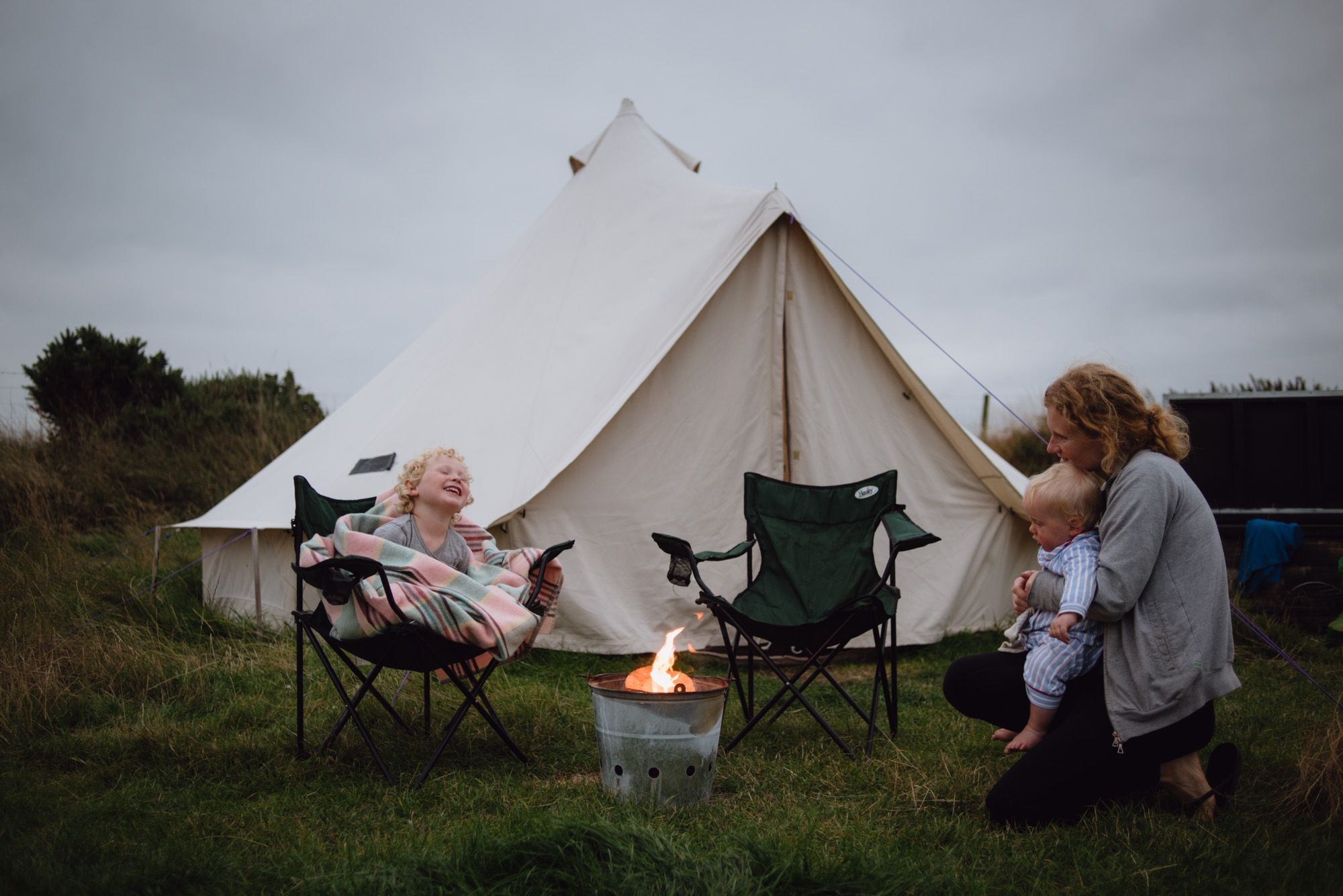 A woman sits around a campfire with her two young children - there's a bell tent in the background.