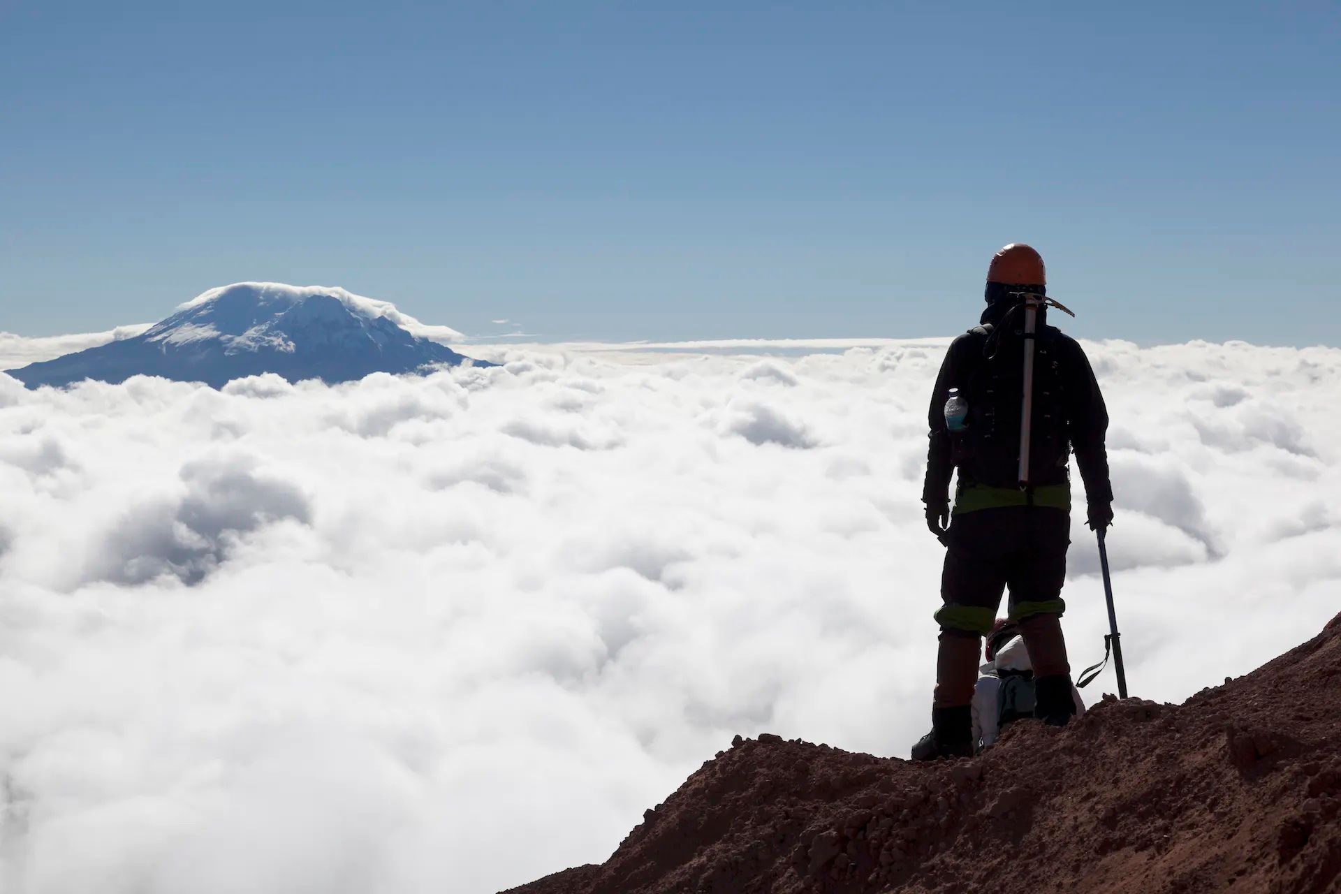 A climber with an ice axe stares across a sea of clouds to a distant mountain summit.