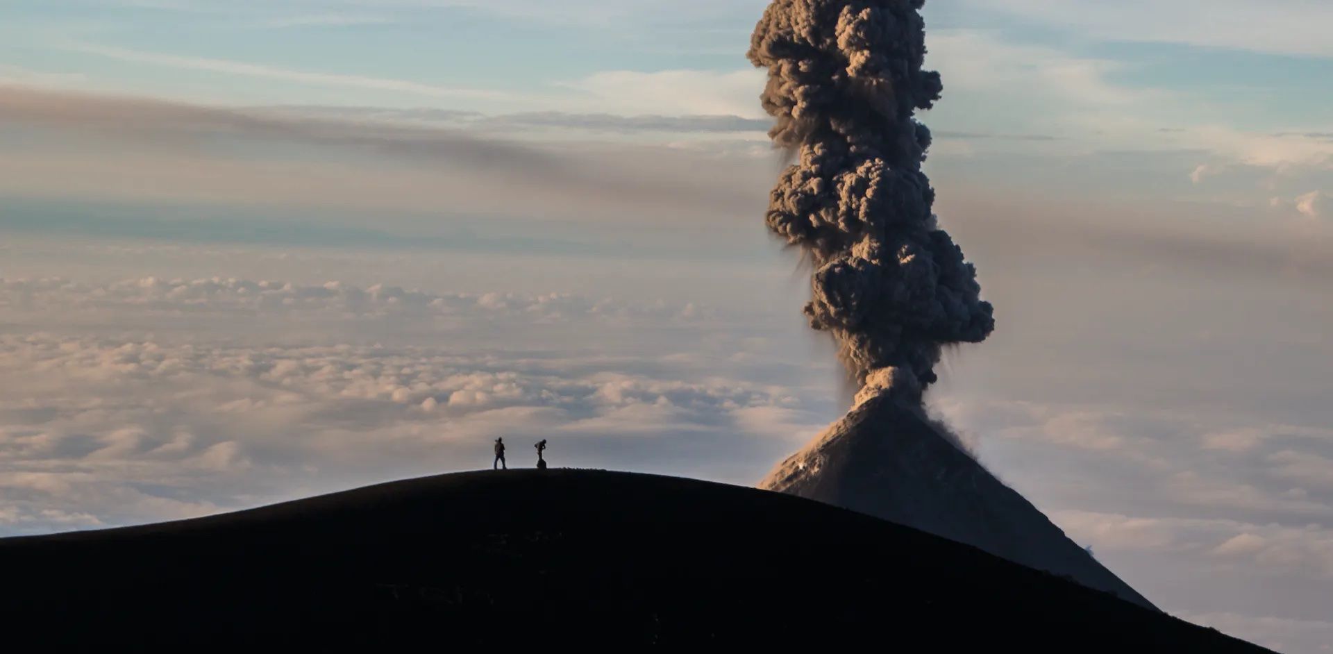 A hiker stands on Tajumalco Volcano in Guatemala, looking at a column of smoke belching out of El Fuego, a nearby volcano