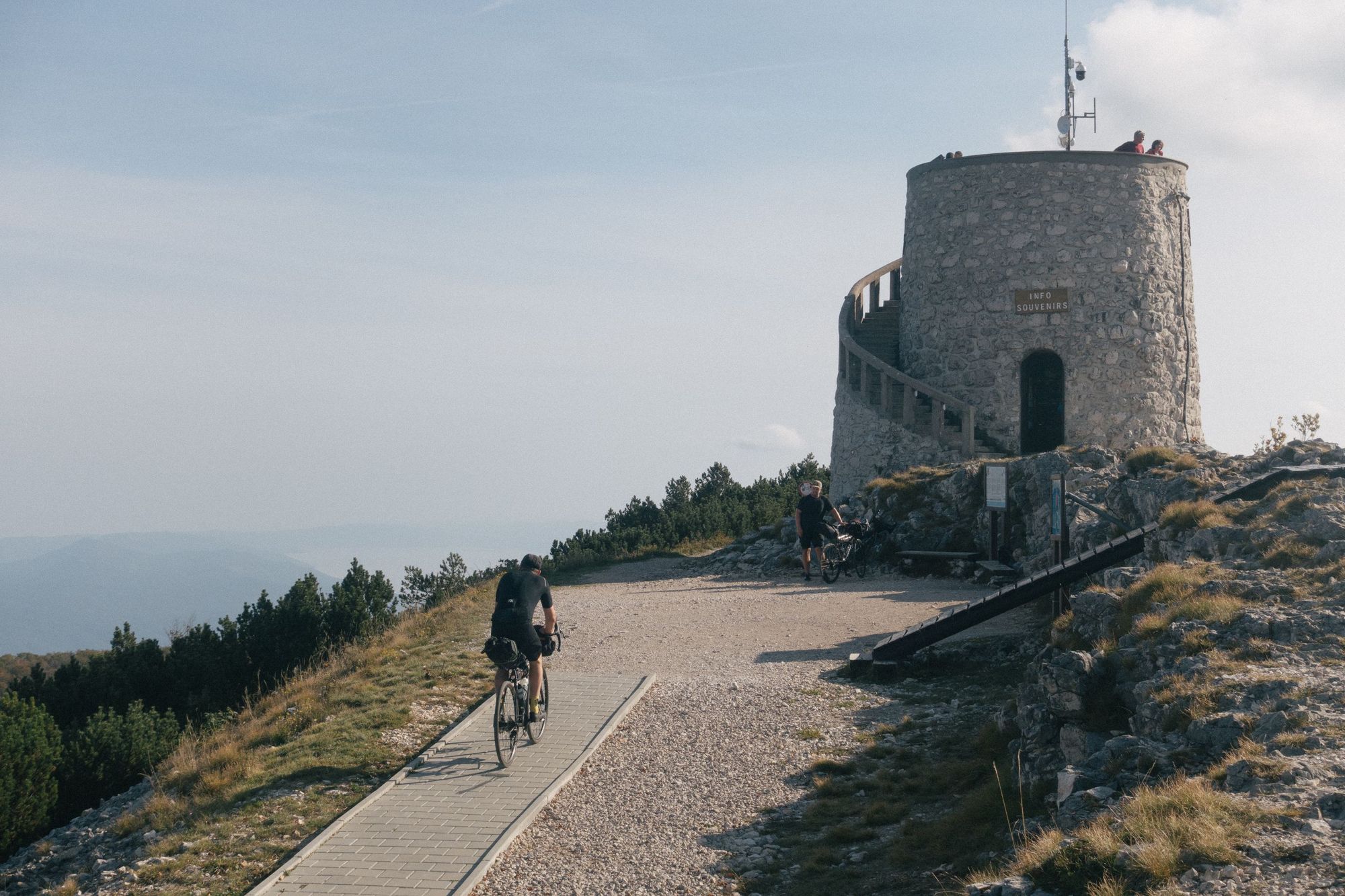 Two cyclists reach the top of a mountain summit in Croatia with a tower of a castle at the peak.