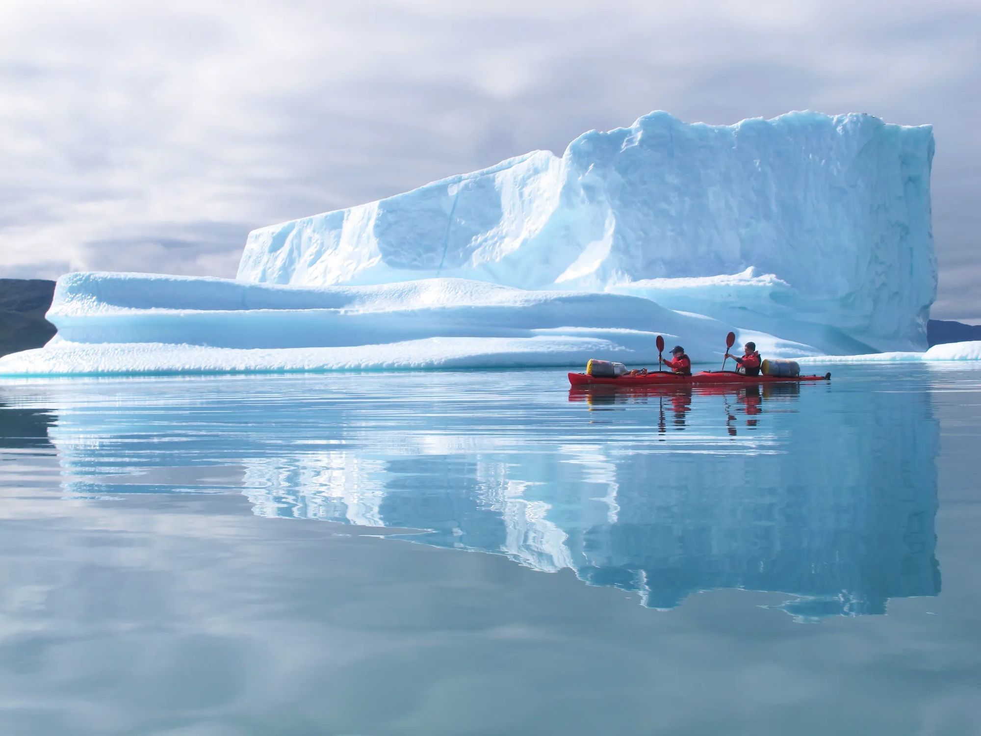Two kayakers paddle past a vast iceberg in Greenland.