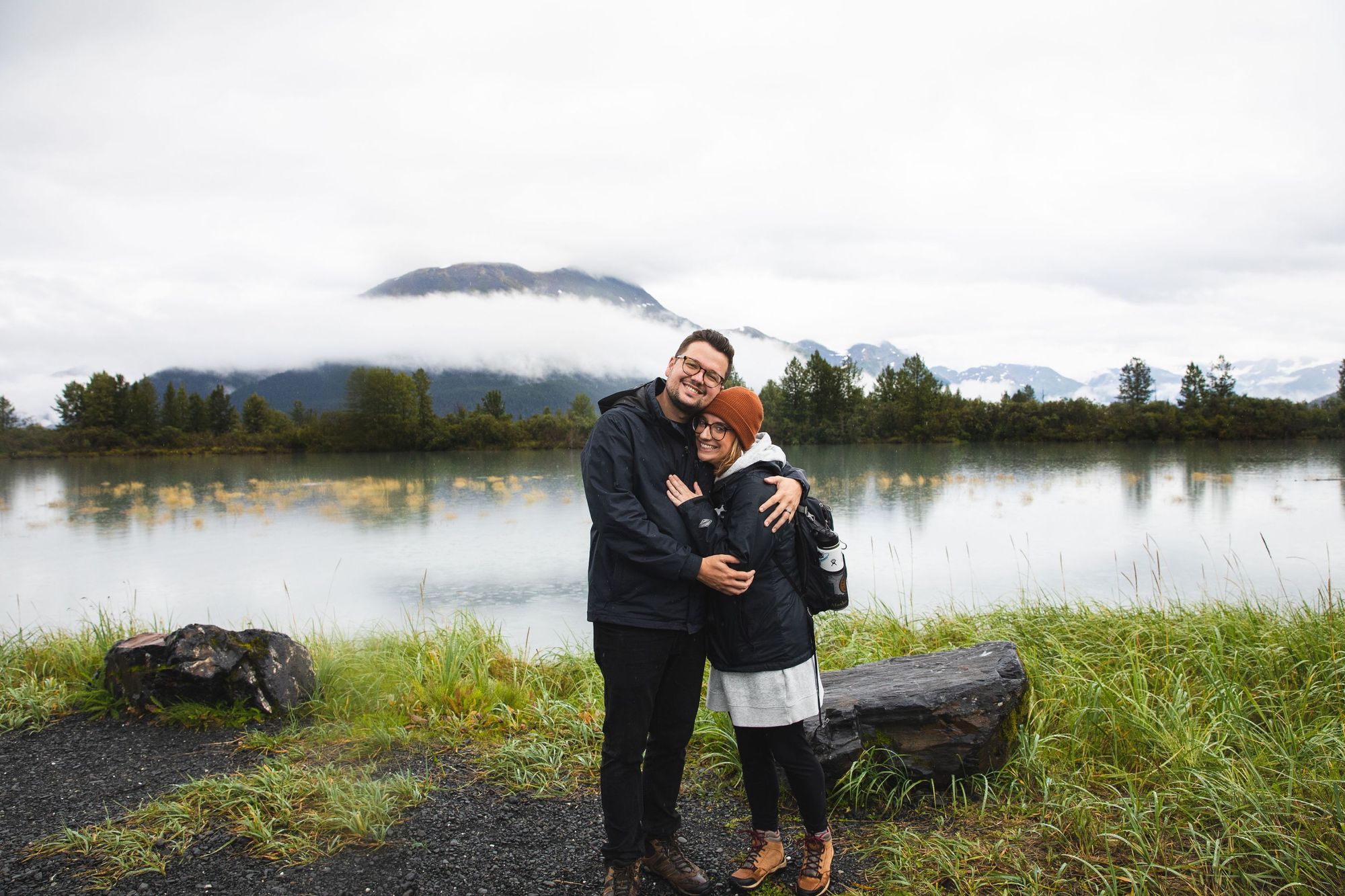 A man and woman pose on a lake shore.