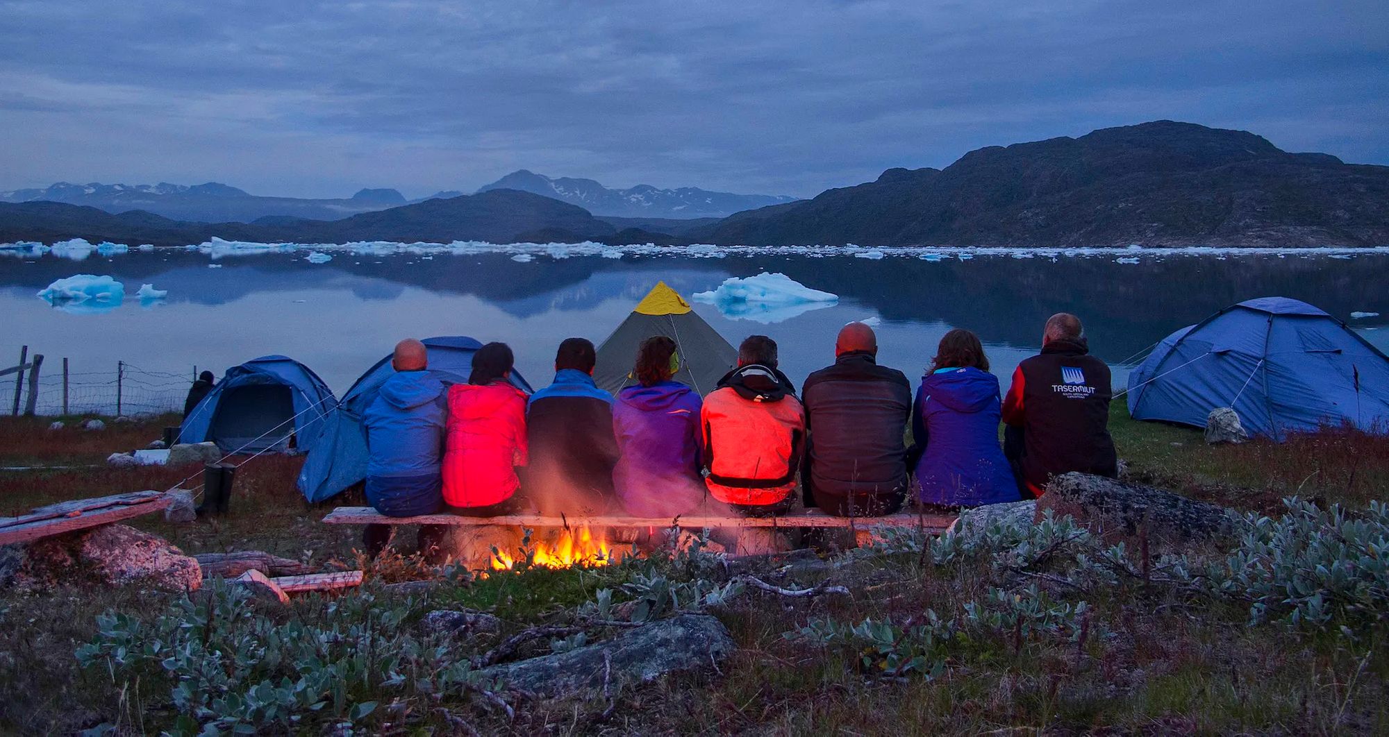 A group of people sit by a campfire at dusk, overlooking an icy fjord in Greenland.