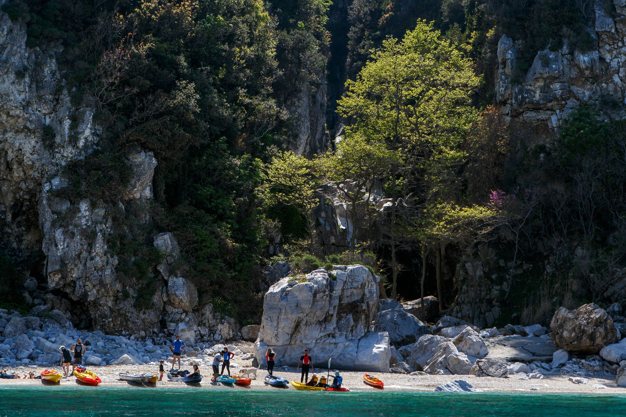A group of kayakers on a beach with a rocky backdrop in the Pelion Peninsula, Greece.