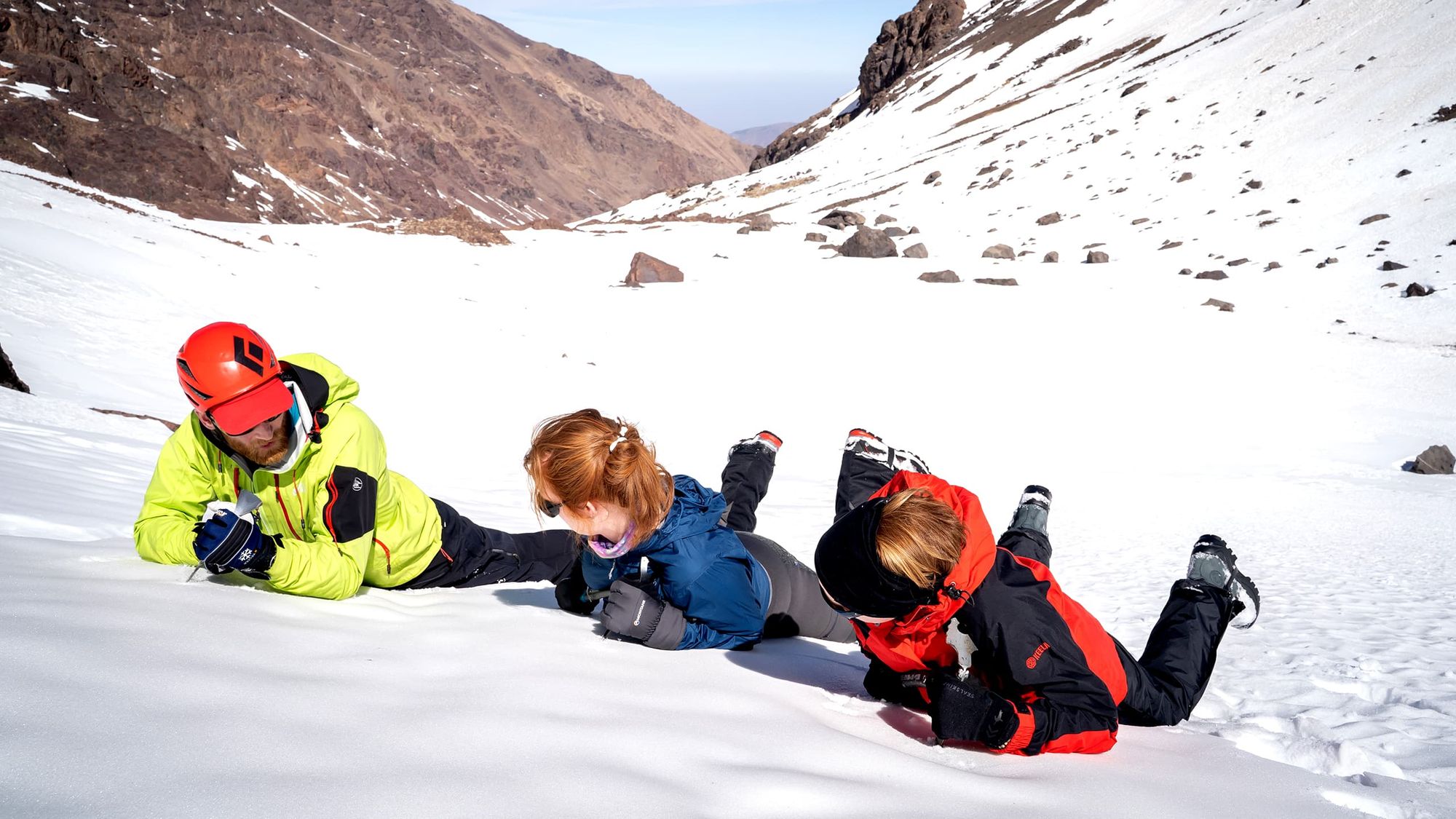 A mountaineering guide demonstrates the 'self-arrest' technique to two women.