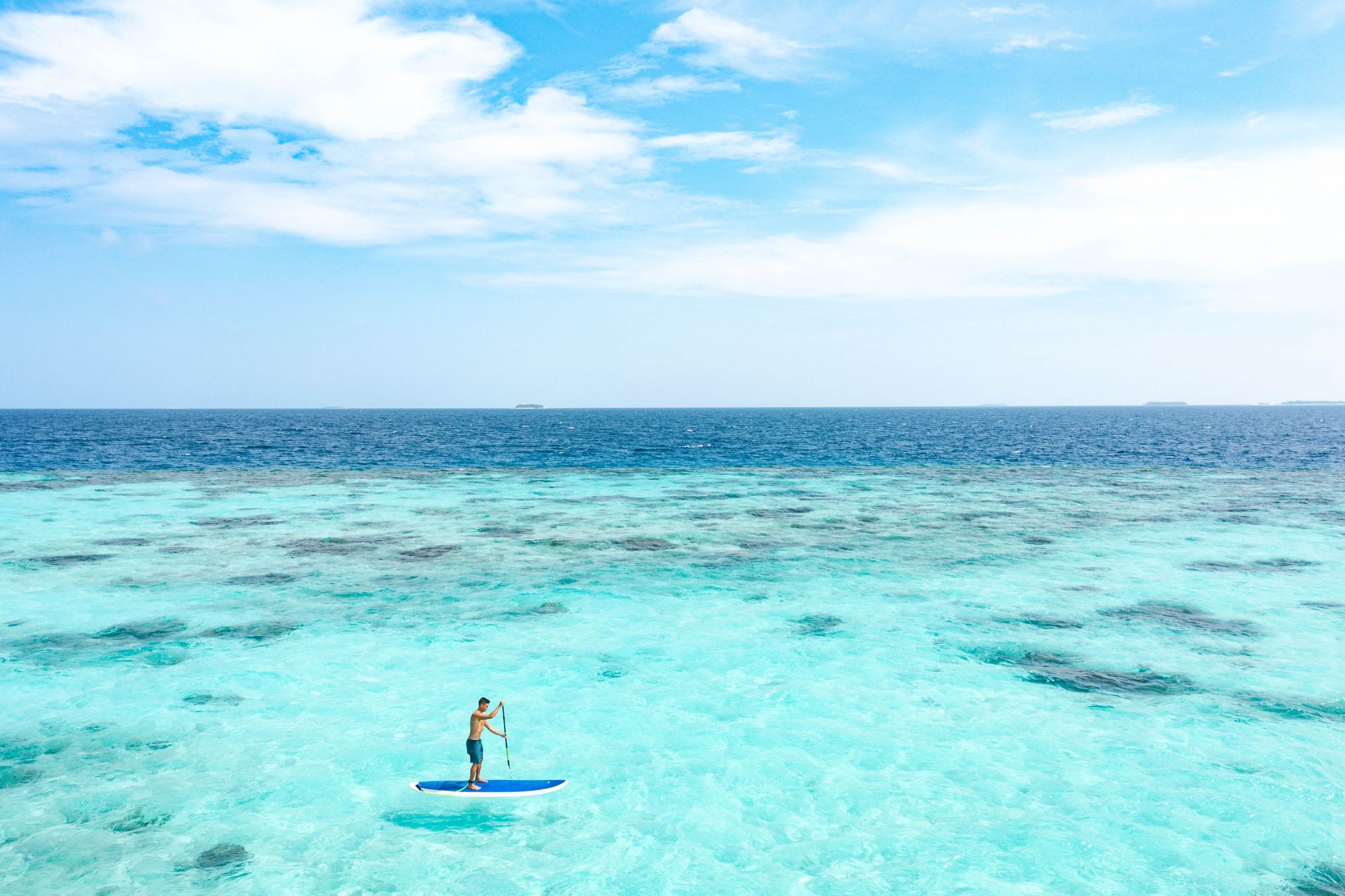 A man stand up paddleboarding in the Maldives, surrounded by turquoise ocean.
