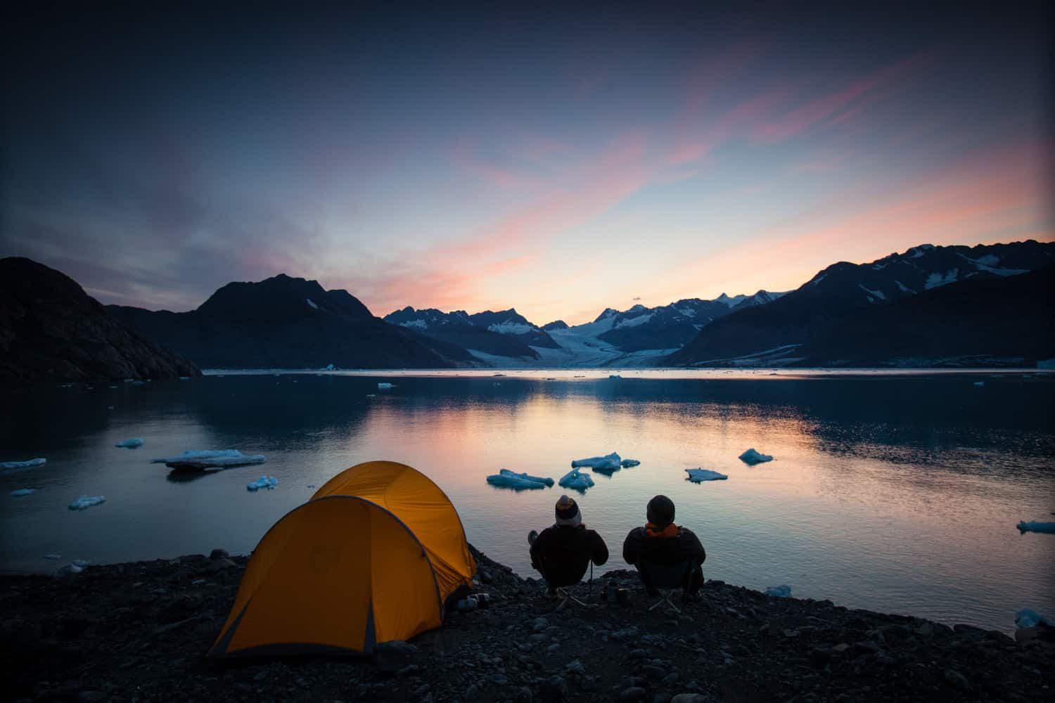 Two campers watch the sun set by a lake in Alaska.