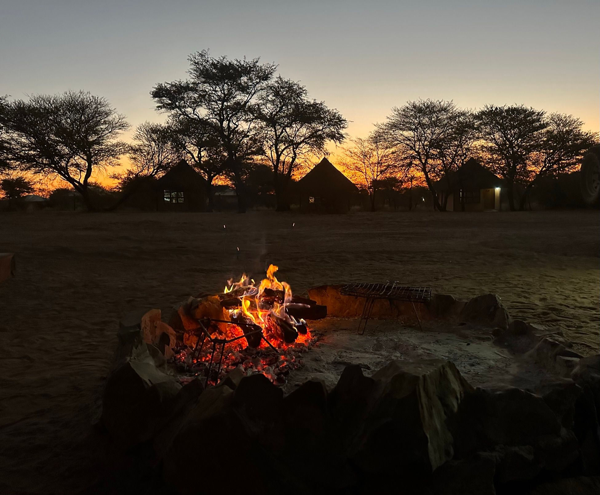 Campfire in the foreground, with a background of tents and trees silhouetted against the sunset.