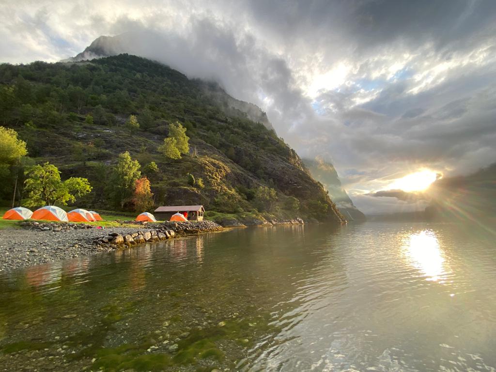 Sun breaking through the clouds over a campsite by the fjord edge.