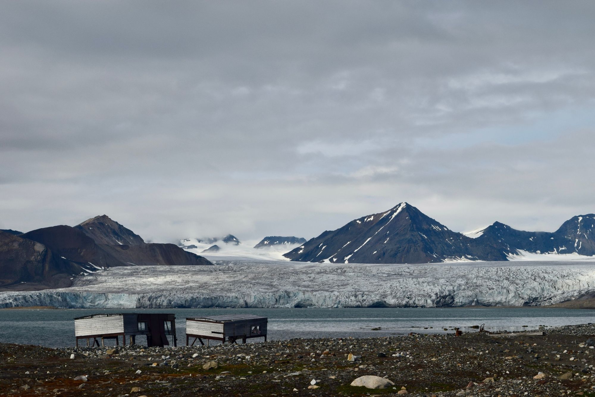 A panorama of the Esmark Glacier and Spitsbergen's snow-dappled mountains, with wooden structures in the foreground.
