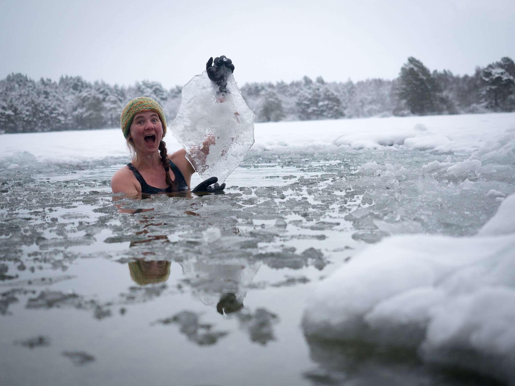 A woman goes ice swimming in a lake surrounded by forest