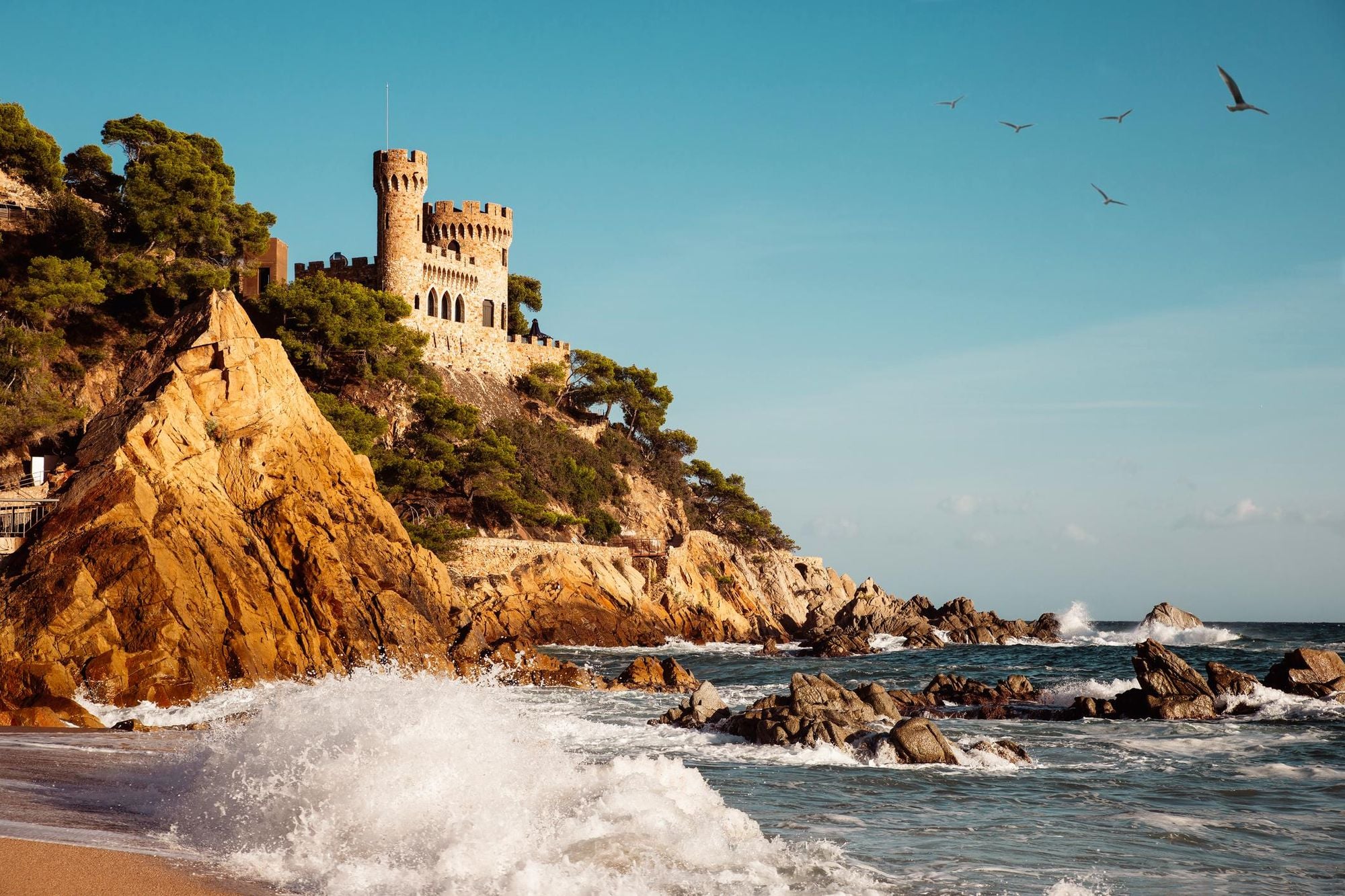 Breaking waves on a beach in Lloret de Mar, Spain with a castle on a rocky cliff above the sea at Costa Brava. Photo: Getty